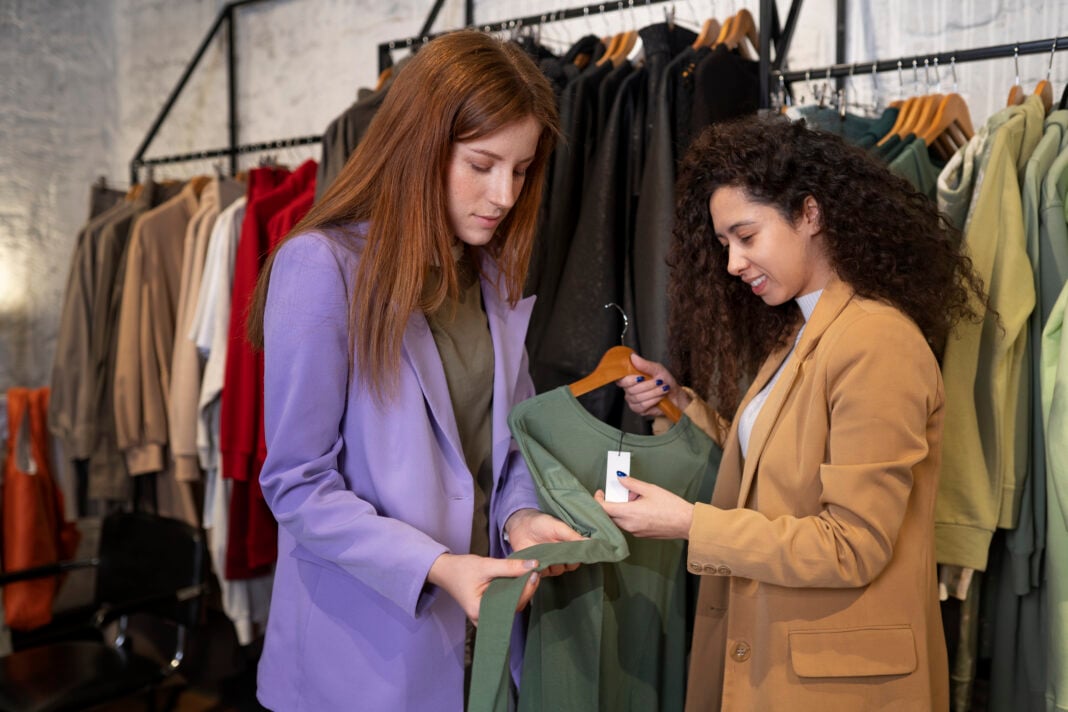 Two women are in a clothing store. One wears a purple jacket, the other a tan coat. They smile while examining a green sweater on a hanger.