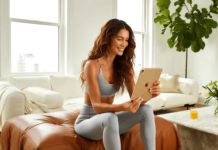 How Sleep Impacts Metabolic Health (And What to Do About It) A woman in a gray workout outfit smiles while using a tablet on a brown ottoman in a bright, modern living room with a large plant and a small table.